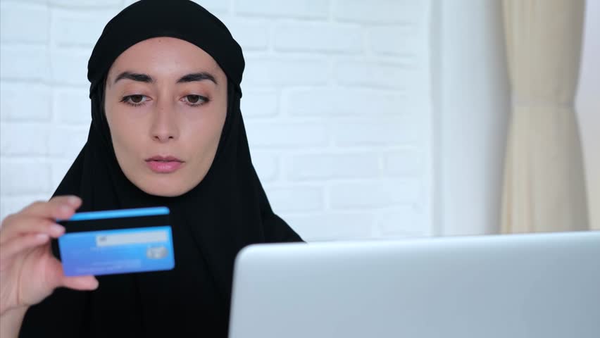 Muslim woman holds card in hands and uses laptop for online shopping or paying for utilities. Woman very carefully drives these boats for payment. Is possible to make any payment without leaving home | Shutterstock HD Video #1111876877