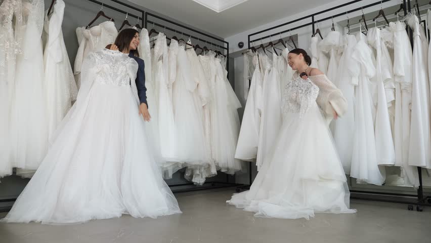 Portrait of two beautiful young women in a wedding salon, they are standing with gorgeous white dresses in their hands in a large bright room. Preparation for the wedding. Royalty-Free Stock Footage #1111881369