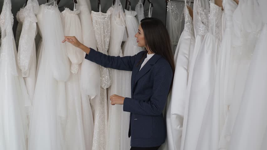 A happy beautiful woman chooses a wedding dress, she walks along the hangers with various dresses in a large modern wedding salon. Preparation for the wedding day. Royalty-Free Stock Footage #1111881377