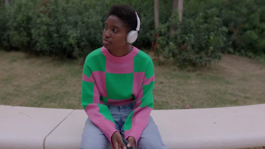 Joyful young african millennial woman enjoying online music while sitting at public park. Black female listening radio podcast or her favorite song while resting outdoors. Technology lifestyle concept | Shutterstock HD Video #1111885349
