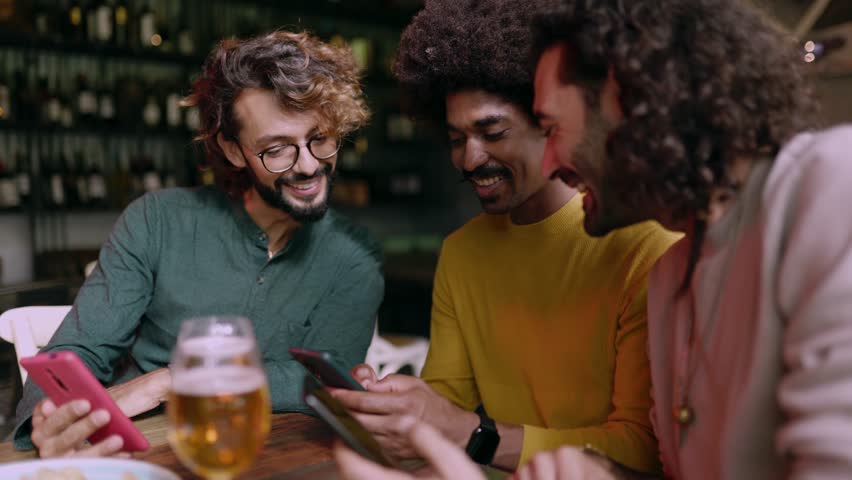 Three diverse young adult friends using mobile phone sitting on table at bar. Technology and male friendship concept. | Shutterstock HD Video #1111885355