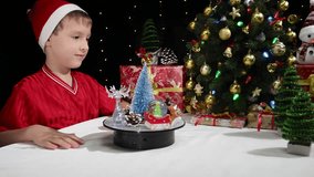 A boy near a Christmas interior watches a small Christmas tree and decorations on a rotating surface