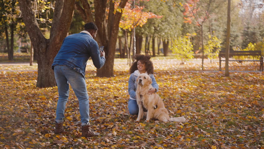 Smiling black girlfriend poses for photo to post on social media with dog made by black boyfriend in park. Young friends have fun in autumn season in nature | Shutterstock HD Video #1111890093