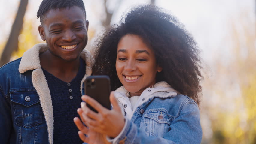 African-American boyfriend looks at photos on phone made together and compliments girlfriend look in autumn time. Couple spends last autumn day outdoors and makes photos | Shutterstock HD Video #1111890103