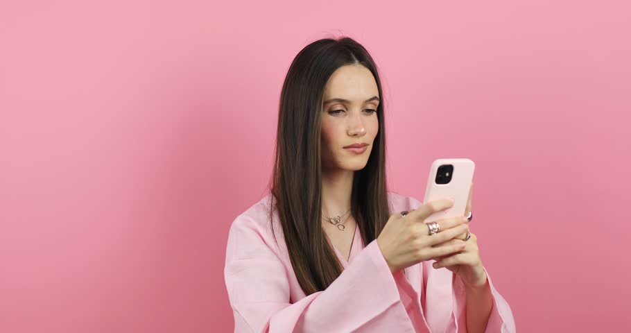 Happy young casual woman in pink dress reading and scrolling social media look happy, point like or dislike say wow standing isolated pink background. | Shutterstock HD Video #1111897631