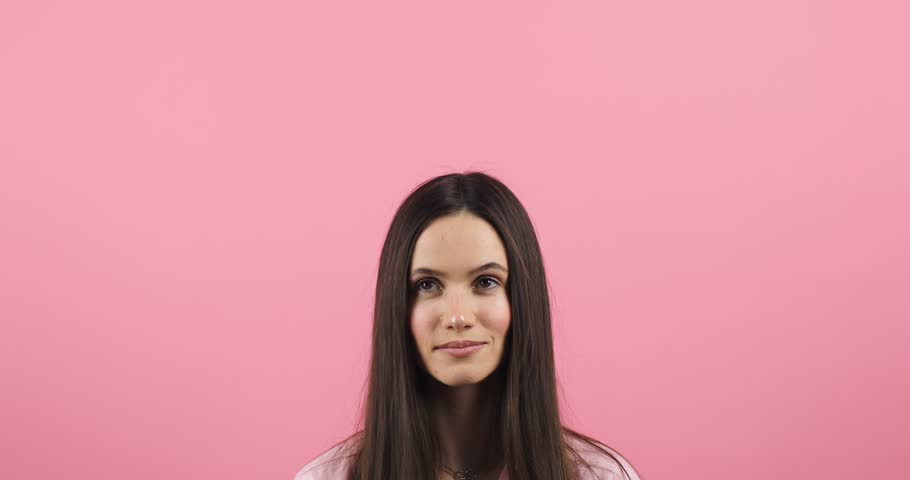 Attractive brunette woman looking at camera and pointing index finger up at copy space for advertising with smile and positive emotions, excited, look up standing isolated pink background. | Shutterstock HD Video #1111898441