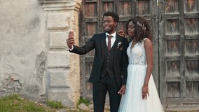 Beautiful African American newly formed young family having video call while standing before castle or church. Loving people smiling at phone camera and showing their wedding rings. Getting married.