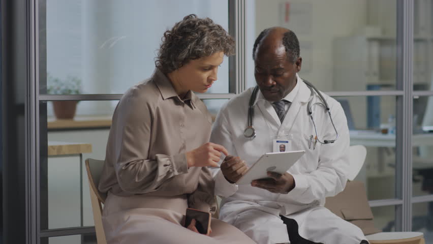 Medium pan shot of black male practitioner with stethoscope and tablet explaining diagnosis to female Caucasian patient sitting in clinic corridor | Shutterstock HD Video #1111900385
