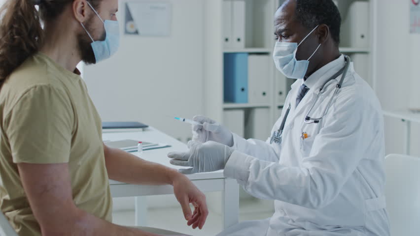 Medium side shot of African American doctor wearing face mask and gloves and making vaccine injection to Caucasian patient in face mask in clinic | Shutterstock HD Video #1111900661