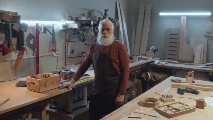 Medium full portrait of senior male carpenter with gray hair wearing apron standing in his workshop looking at camera | Shutterstock HD Video #1111901199