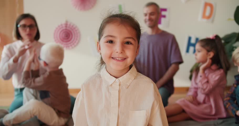 Portrait: happy little girl with brown eyes in a white shirt looks at the camera and smiles at her first lesson in the pre-school club. The little girl sits at her first lesson in the school Royalty-Free Stock Footage #1111901483