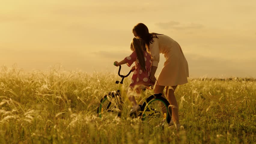 Caring mother teaches daughter riding bicycle on field in summer. Mother supports child riding bicycle in meadow on vacation. Happy mother with daughter riding bike spends summer weekend at sunset | Shutterstock HD Video #1111902121