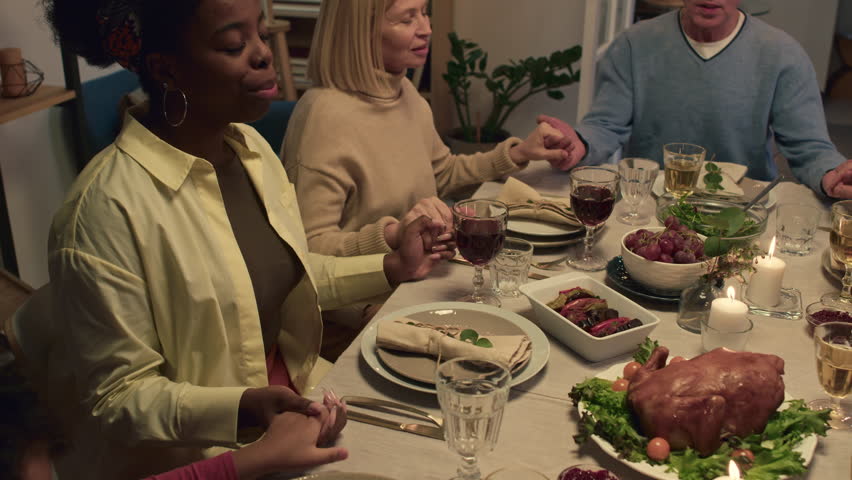Members of multi-ethnic family praying before festive dinner sitting by served table with dishes and wine in cozy living room | Shutterstock HD Video #1111902451