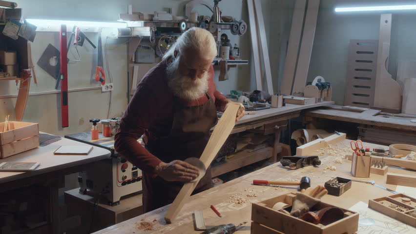 Medium full shot of senior Caucasian carpenter with bushy gray beard smoothing out wooden stem using sandpaper | Shutterstock HD Video #1111902863