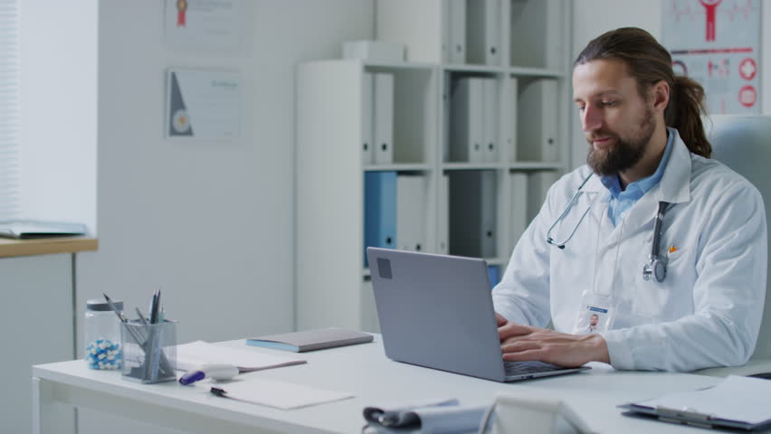 Medium pan shot of Caucasian male doctor in medical outfit working on laptop in hospital office | Shutterstock HD Video #1111903105