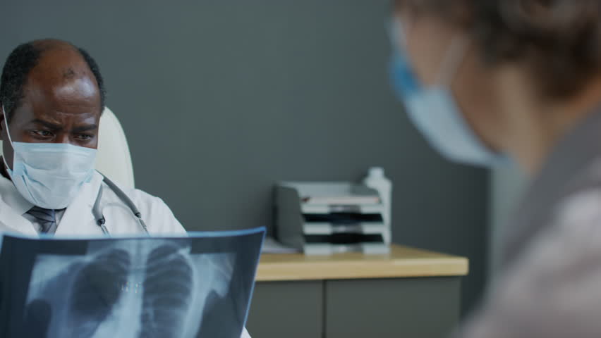 Close up panning shot of black male physician wearing face mask examining chest x-ray and discussing results with patient in hospital | Shutterstock HD Video #1111905113