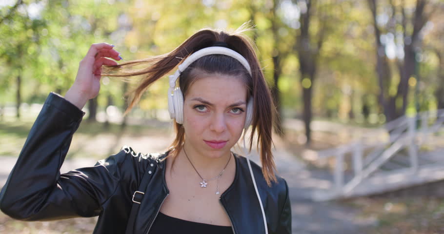 Fashionable young lady in the park portrait. Beautiful girl with headphones looking at camera. Slow motion recorded at 120fps.