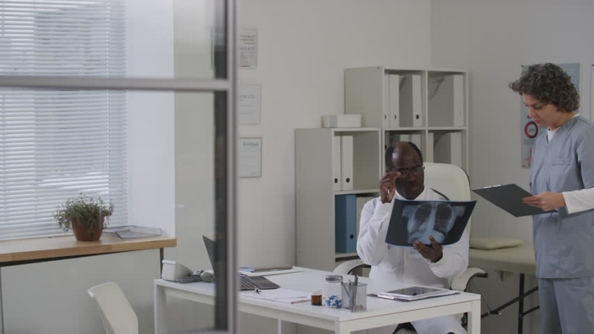 Medium pan shot of female Caucasian medical professional consulting with experienced black male doctor in glasses about chest x-ray scan in medical office | Shutterstock HD Video #1111907441