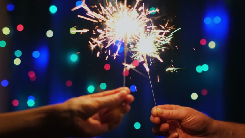 Burning bengal lights close-up. Hands holding sparklers on festive bokeh background with blinking garland in the dark. Winter holidays concept. Merry Christmas and Happy New Year celebration.