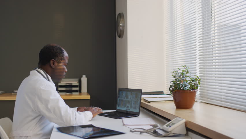 Medium side shot of black male doctor in medical uniform participating in conference call with colleagues using laptop during workday in clinic | Shutterstock HD Video #1111908269
