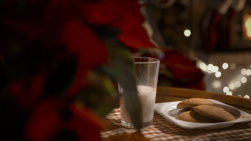 Santa Claus putting Christmas presents in socks over the fireplace, eating ginger cookies and drinking milk on Christmas night in the decorated living room with Christmas tree