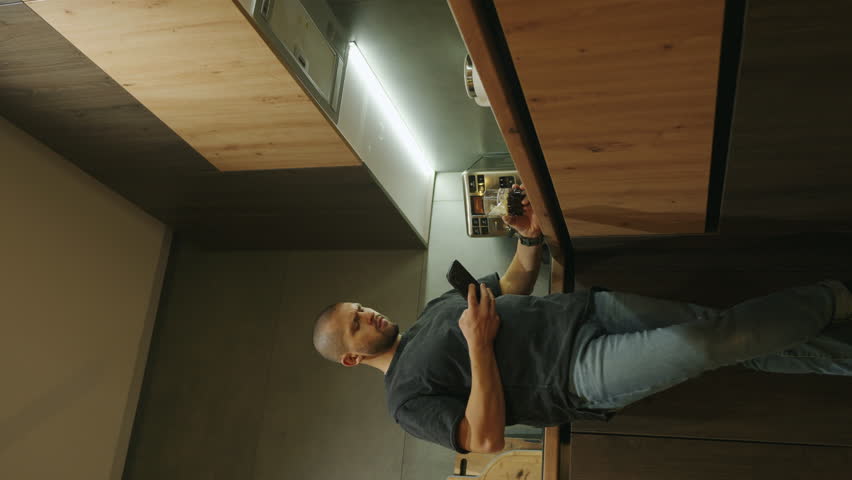 Fron view of an adult man relaxing at home in kitchen holding glass of drink using mobile phone. Carefree day. Handsome young man in casual wear drinking wine.