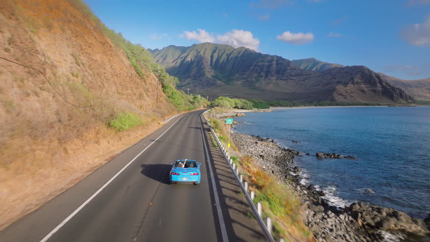 Summer vacation adventure tourism. Aerial view blue convertible car driving by coastal road along dramatic rocky shore towards volcanic mountains on Oahu island. Happy young couple traveling by Hawaii