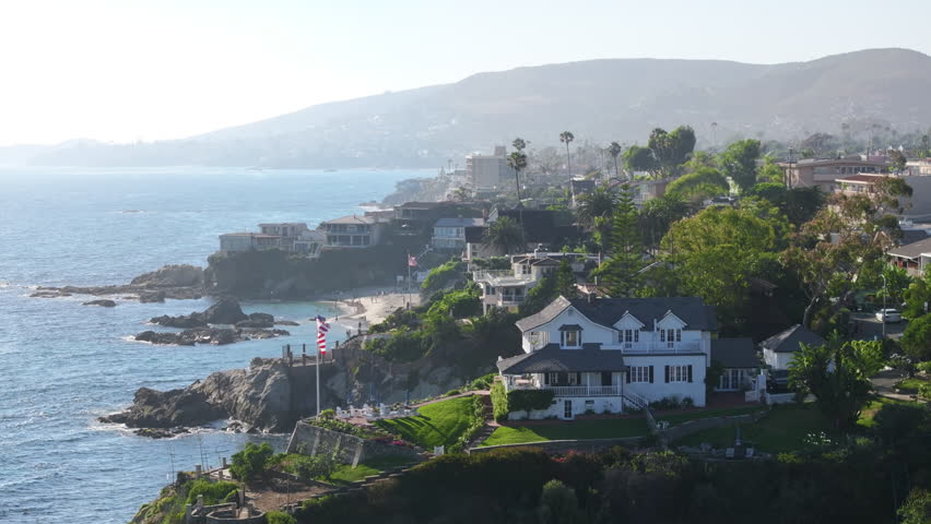 Birds eye view of picturesque Laguna beach coastline, Orange County, California, USA. Seaside resort at the Pacific Ocean. Residential houses with mountains landscape. American flag waving by wind