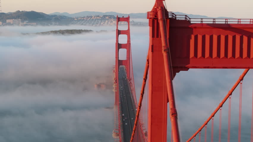 Impressive aerial of cinematic Golden Gate bridge in sunrise light. Parallax dolly shot with close up view on red tower soaring above dense fog covering ocean. USA landmark, tourism copy background 4K