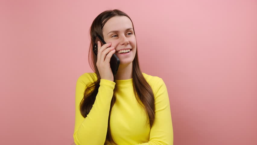 Cheerful cute young woman talking with friend on cell phone, looking away and smiling joyfully, having pleasant conversation, wearing yellow sweater, posing isolated on pink background wall in studio | Shutterstock HD Video #1111920589