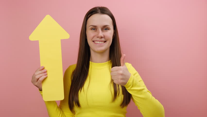 Satisfied young brunette woman holding yellow arrow pointing up looking at camera with smile, growth and increase concept, wearing sweater, posing isolated over pink color background wall in studio | Shutterstock HD Video #1111922549