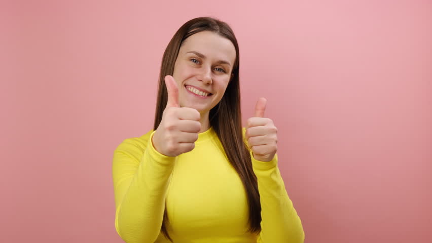 Attractive young woman showing double thumbs up, gesturing like to camera, girl smiling broadly, expressing approval, positive feedback, wearing yellow sweater, isolated on pink studio background | Shutterstock HD Video #1111922551