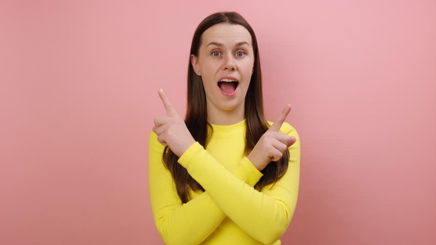 Portrait of pleasantly surprised cute young woman standing with crossed hands and pointing to copy space on both sides, wearing yellow sweater, posing isolated on pink color background wall in studio | Shutterstock HD Video #1111922689