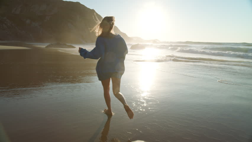 Happy young woman in oversized sweater run barefoot on beach in ocean surf waves. Cinematic moment of happiness and joy on warm summer evening | Shutterstock HD Video #1111923863