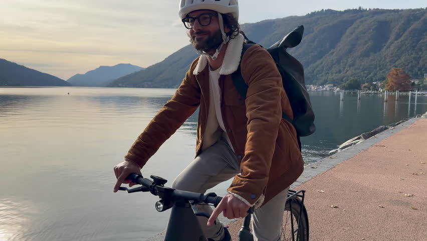 Man rides commuter city electric bike along lakefront walkway, dressed in fall attire. He admires open lake view  Royalty-Free Stock Footage #1111925057