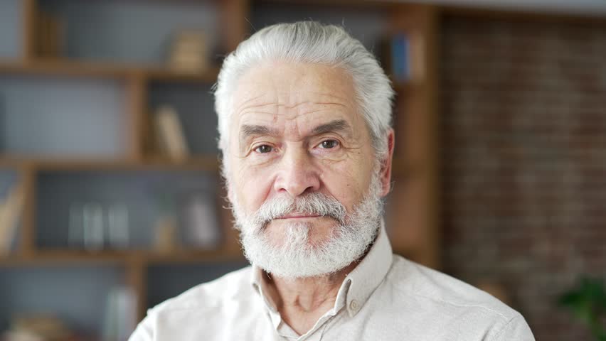 Close up portrait of a happy gray haired old man standing in the living room at home. Handsome senior retired male posing looking at camera. Headshot of confident smiling bearded grandfather indoor