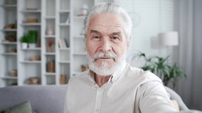 POV Elderly senior gray haired man talking on a video call taking a selfie using a smartphone sitting on sofa in living room at home. A smiling positive bearded adult mature pensioner has online chat