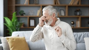Elderly senior gray haired man suffering from eye fatigue while sitting on sofa in living room at home. Overtired adult mature male takes off his glasses and massages bridge of his nose with his hand - Powered by Shutterstock - Get 15% off with code: PIKWIZARD15
