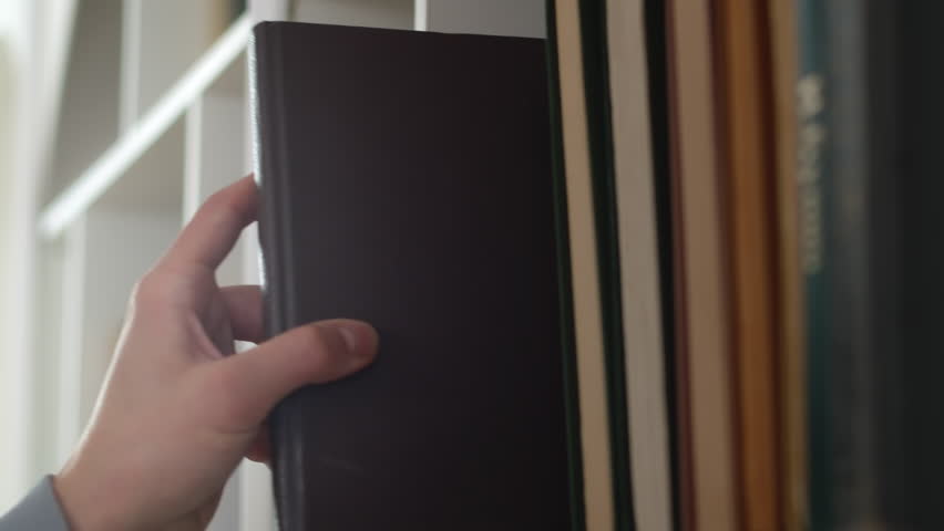 Close-up of a hand carefully taking a book out of big range staying in row in a shelf of a white bookcase at home. Concept of curiosity and thirst for knowledge