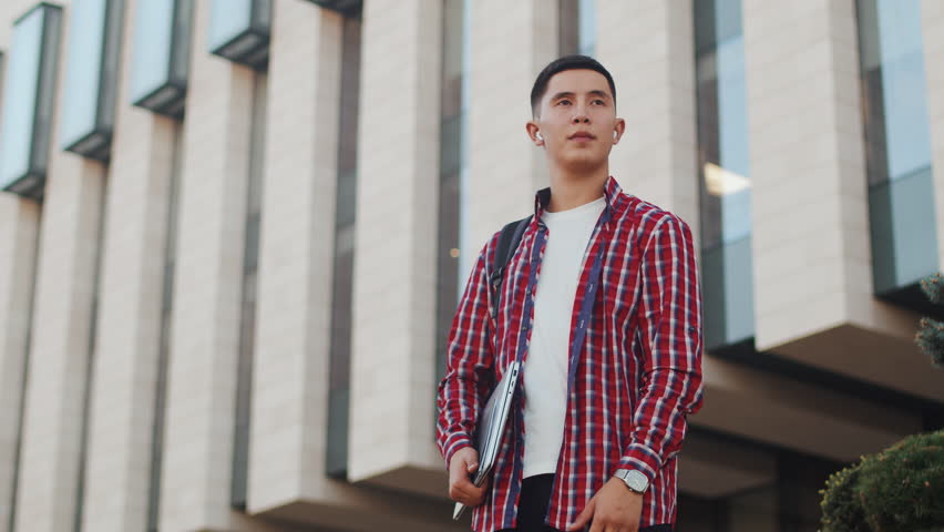 Young asian guy with a backpack and a laptop in his hands takes a look around and then looks at the camera and smiles outdoors with a building in the backdrop. Concept of modern remote work with a | Shutterstock HD Video #1111926851