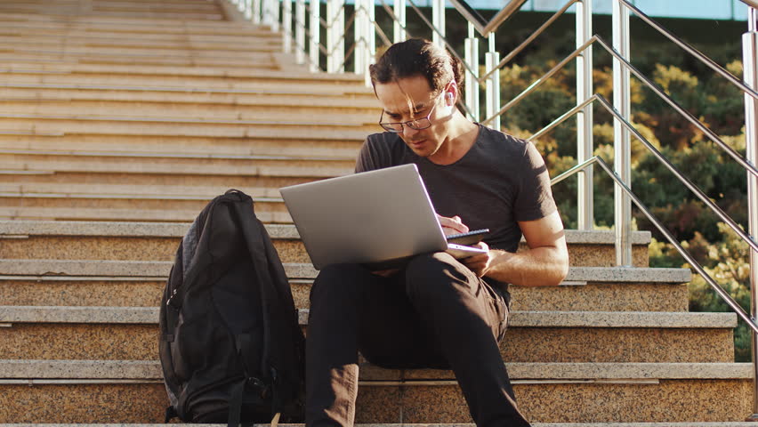 Young asian man in glasses sitting on the stairs with a laptop and backpack and writing in the notebook with a focused face. Concept of modern remote work with a flexible schedule | Shutterstock HD Video #1111926853