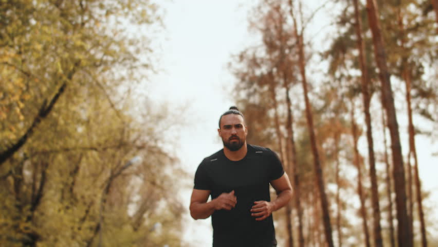 Young muscular jogger with a focused face calmly running on the path of a city park on a sunny day, front view. Concept of modern outdoor sports training in a casual urban life | Shutterstock HD Video #1111926861