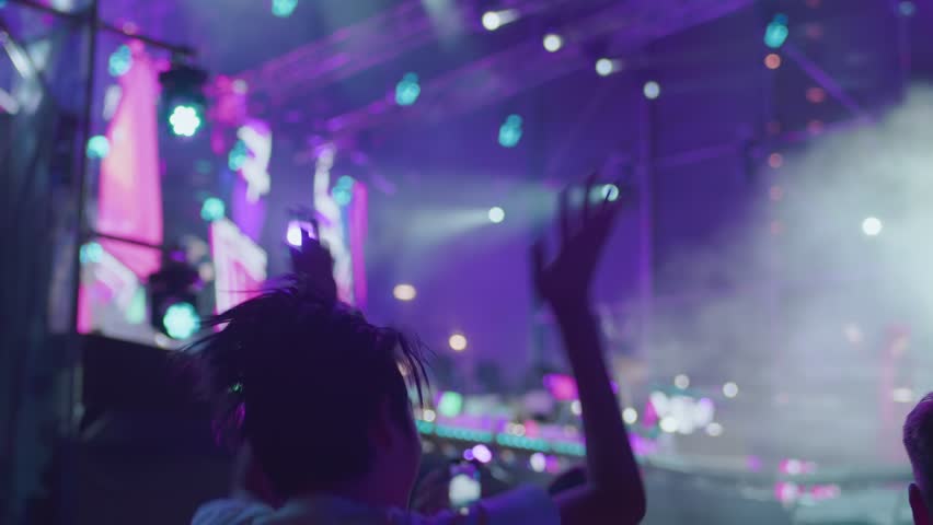 A rear view of a young girl joyfully jumping and clapping her hands at a music concert, festival, event. In the background, the stage is brightly illuminated by floodlights, iridescent lights | Shutterstock HD Video #1111927045