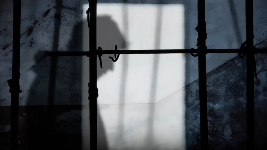 The dark silhouette of a man: walking, stopping to hide his face under the hood, going on incognito. Seen from the cracks on the gate of a prison cell, sunlight projecting a shadow on the wall.
 | Shutterstock HD Video #1111927713