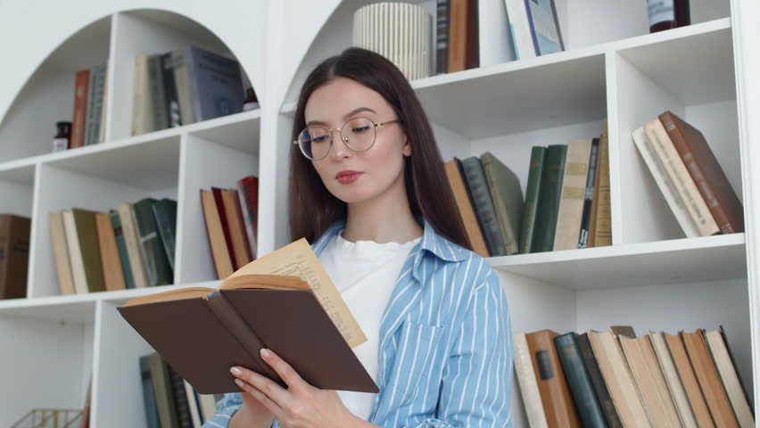 Young attractive woman in glasses reading the book with focused face and flipping pages with the backdrop of white bookcases with many books on the bookshelves. Concept of curiosity and thirst for | Shutterstock HD Video #1111928217