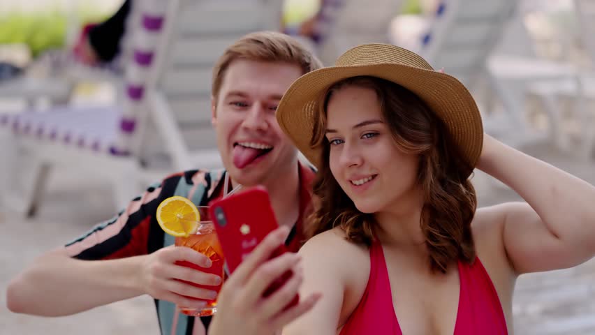 Happy man and woman photographing vacation at poolside. Girlfriend with boyfriend show v sign when making a selfie on summer pool party outdoors. Funny man drink cocktail send greetings with lady | Shutterstock HD Video #1111930391