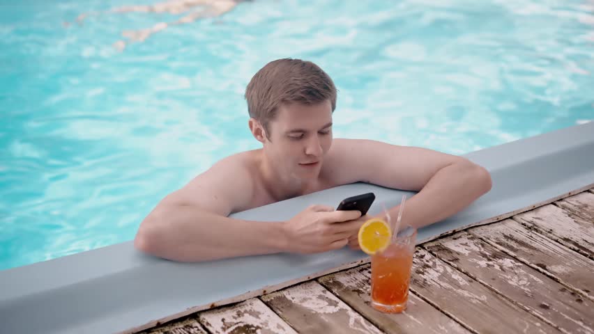 Young handsome man using smartphone while sunbathing in swimming pool and drinking cocktail. Happy male tourist chilling texting phone poolside outdoors. Chill in cool blue water | Shutterstock HD Video #1111930395