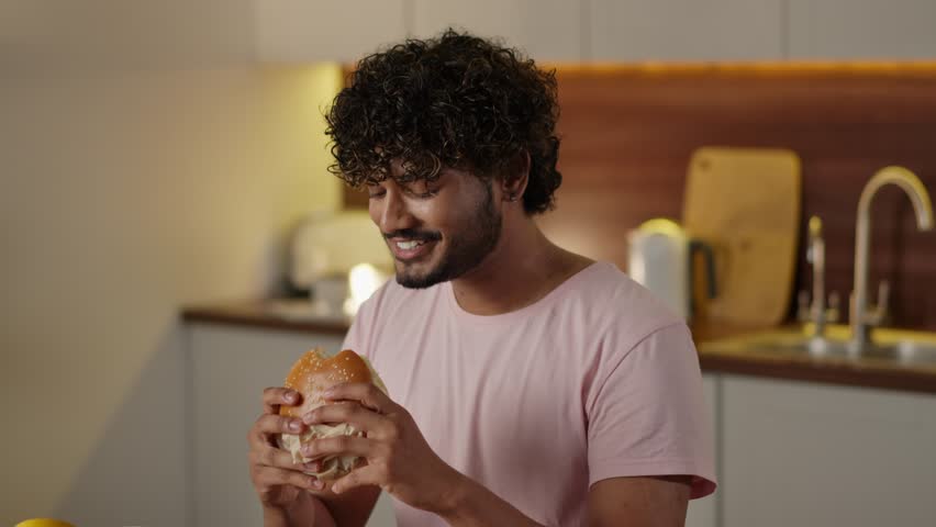 Concept of unhealthy food delivery. Hindu guy enjoying his hamburger. Young man with kinky hair biting tasty burger in kitchen. Male student eating big sandwich Royalty-Free Stock Footage #1111930397