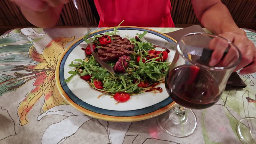 Woman eating a vibrant feast of medium-rare steak salad on a plate, paired with a glass of rich red wine, set against a backdrop of a colorful floral tablecloth. Served with salad and red wine | Shutterstock HD Video #1111931285
