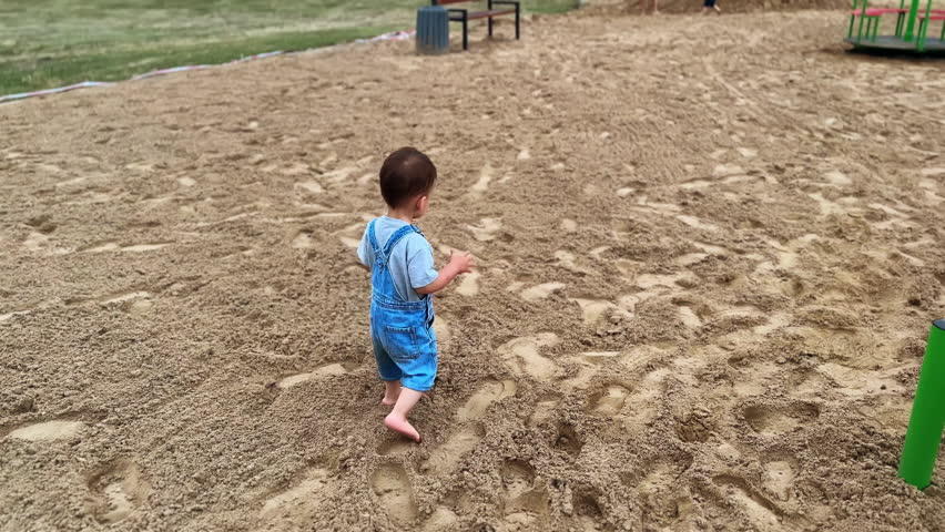 Following the little kid in jeans romper walking barefoot by the sandy ground. Lovely toddler heads to the merry-go-round.
